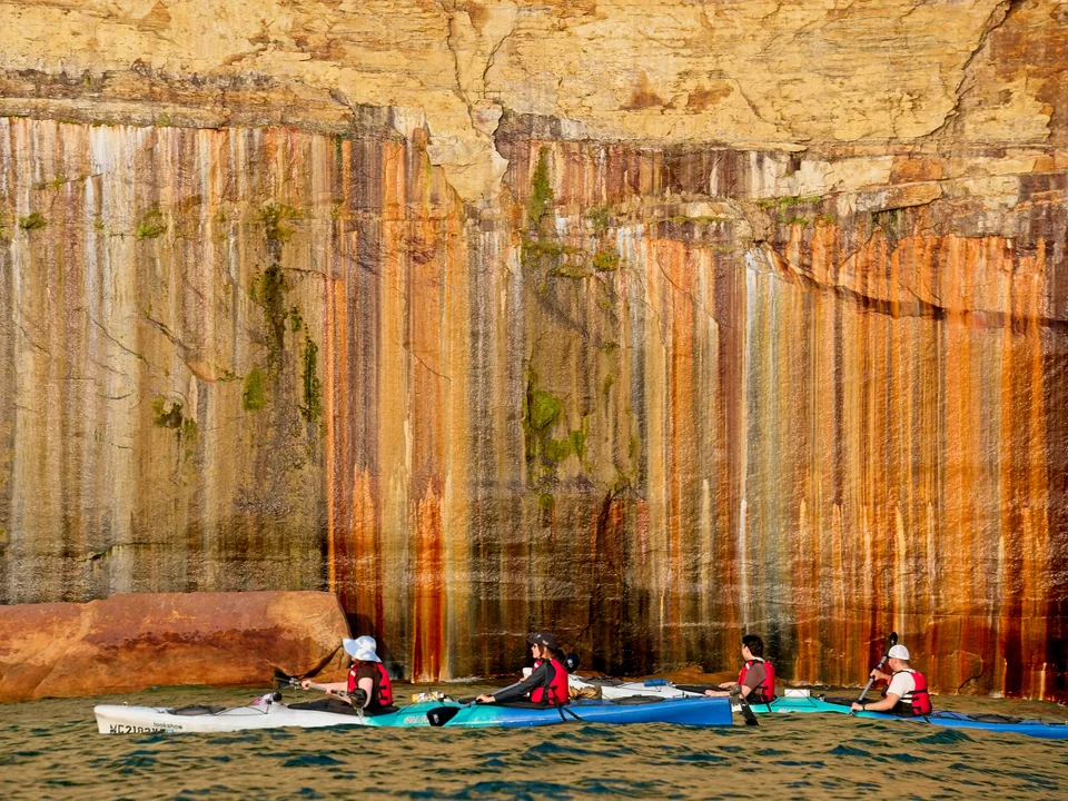 Kayakers trace vivid orange, green, and black mineral bands painted down the cliff face.