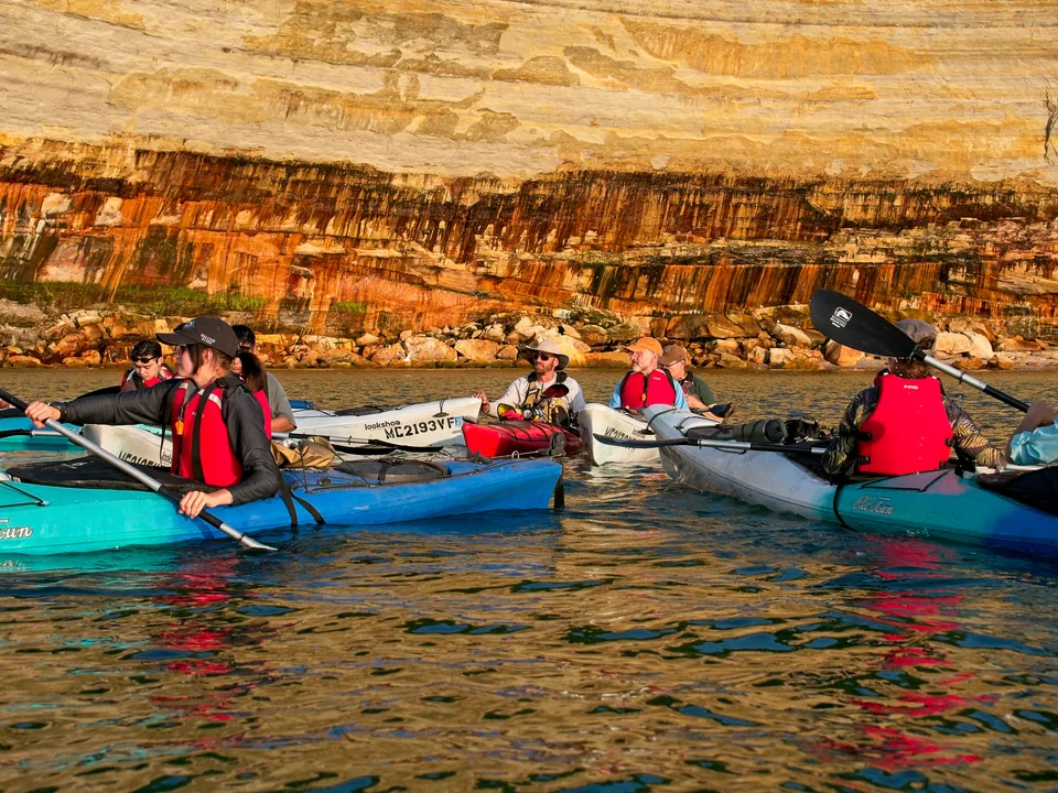 The tour pauses together beneath layered sandstone, bright boats gathered against the rock.