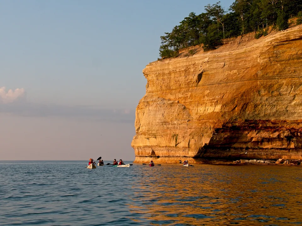 A line of tiny boats rounds a massive sunlit headland on Lake Superior.